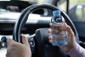 asiatische Fahrerin, die eine Flasche zum Trinken von Wasser hält, während sie ein Auto fährt. Wärmflasche aus Kunststoff verursacht Feuer. foto