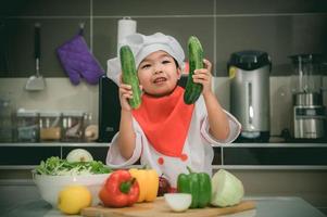 süß asiatisch Mädchen tragen Koch Uniform mit ein Menge von vegatable auf das Tabelle im das Küche Zimmer, machen Essen zum Essen Abendessen, lustig Zeit zum Kinder foto