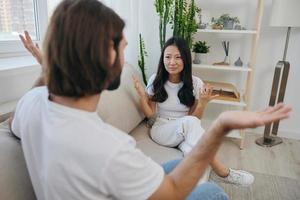 ein Mann und ein Frau sitzen auf das Couch beim Zuhause im Weiß T-Shirts und schreien kommunizieren skandalös tun nicht verstehen jeder andere. ein Streit im das Familie von zwei Ehepartner und Aggression, traurig Emotionen foto