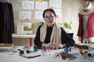 asiatische reife weibliche modedesignerin sitzt am tisch, schaut in die kamera und im studio, arbeitet mit kreativen und näharbeiten für die kleiderdesign-kollektion, professioneller boutique-schneider-sme-unternehmer. foto