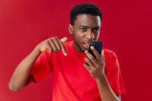 Mann von afrikanisch Aussehen im ein rot T-Shirt mit ein Telefon im das Hände von Kommunikation Technologie foto