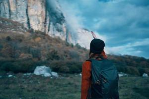 ein Reisender mit ein Rucksack und im ein rot Sweatshirt zeigt an das Berge mit ihr Hand und das Blau Himmel Wolken foto