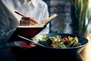 Frau mit Notizblock in der Nähe von das Fenster und Salat im ein Teller Tomaten frisch Gemüse foto