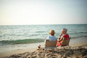 ein Paar von Alten Sitzung im Stühle beim das Strand Aufpassen das Sonne und das Meer auf ihr Sommer- Ferien und Sie Lächeln und genießen ihr Urlaub. foto