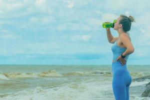 schön blond Sportlerin im ein Blau Trainingsanzug während Fitness auf das Strand, Getränke Wasser von ein Shaker, Kopieren Raum foto