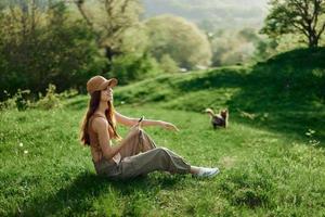 ein Frau Sitzung auf das Grün Gras im ein Park im Sommer- gegen ein Landschaft von Bäume, das Arbeit von ein Freiberufler und Blogger foto