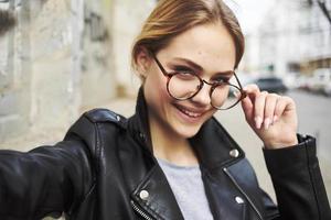 Porträt von ein glücklich Frau im Brille und im ein Leder Jacke draußen lächelnd Modell- foto