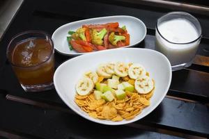 kontinental Frühstück Cornflakes mit Früchte, Milch und Tee und etwas von anbraten Gemüse auf schwarz Holz Tisch. oben Sicht. foto