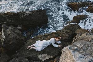 barfuß Frau im lange Weiß Kleid nass Haar Lügen auf ein felsig Cliff Ferien Konzept foto