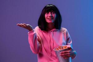 glücklich schockiert genial brünett Frau im Rosa Kapuzenpullover mit Popcorn halt Palme oben posieren isoliert im Blau violett Farbe Licht Studio Hintergrund. Neon- Party Cyberpunk Kino Konzept. Kopieren Raum foto