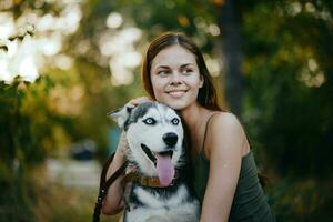 ein Frau mit ein heiser Rasse Hund lächelt und liebevoll Schlaganfälle ihr Geliebte Hund während Gehen im Natur im das Park im Herbst gegen das Hintergrund von Sonnenuntergang foto