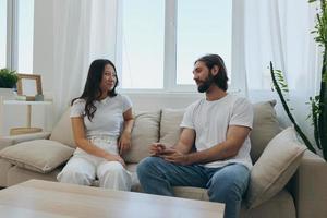 ein Mann und ein Frau Sitzung beim Zuhause auf das Couch im Weiß stilvoll T-Shirts und chatten fröhlich lächelnd und Lachen beim heim. männlich und weiblich Freundschaft foto