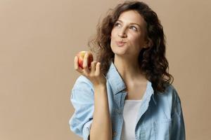 komisch glücklich süß lockig schön weiblich im Jeans beiläufig Hemd Kauen genießen Apfel posieren isoliert auf Über Beige Pastell- Hintergrund. gesund Lebensmittel. natürlich umweltfreundlich Produkte Konzept. Kopieren Raum foto