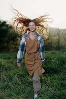 Frau Gehen auf das Gras im Natur lächelnd im Bauernhof Kleider mit schön Haar fliegend im das Wind glücklich im das Strahlen von Sommer- Sonnenuntergang Licht foto