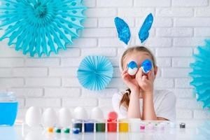 das wenig Mädchen versteckt sich ihr Augen hinter Sie mit Blau Ostern Eier. ein Kind mit Hase Ohren ist genießen Ostern. Zuhause Dekoration im Blau Töne. foto