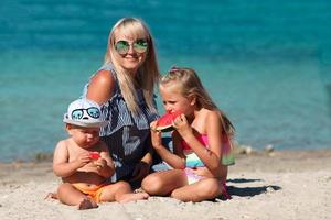 Mama und Kinder sind Sitzung auf das Strand in der Nähe von das Meer und Essen Wassermelone. Urlaub. foto