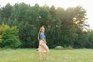 jung kaukasisch Frau oder Mädchen im ein Sommer- Kleid und ein Denim Hemd ist Gehen auf das Gras, halten Schuhe im ihr Hände, entspannend draußen das Stadt auf ein Wochenende im Sommer- foto