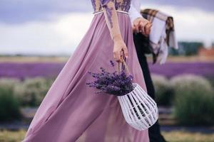 schöne junge Frau in einem blauen Kleid hält einen Blumenstrauß Lavendel in einem Korb beim Gehen im Freien durch ein Weizenfeld bei Sonnenuntergang im Sommer. Provence, Frankreich. getöntes Bild mit Kopierplatz