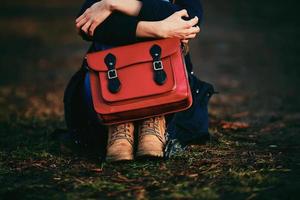 stilvolles junges Mädchen in braunen Schuhen und einem warmen Mantel, der im Park mit einer roten Tasche sitzt. foto