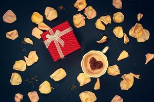 eben legen. rot Box mit ein Bogen, ein Kaffee Becher mit Sahne und Zimt im das bilden von ein Herz mit Rosa und Gelb Blume Blütenblätter auf ein dunkel Blau Hintergrund. Vignettierung. foto