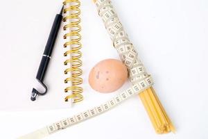 Bündel von roh Spaghetti gebunden mit Messung Band, Hähnchen Ei und Notizbuch zum Schreiben mit schwarz Stift auf Weiß Hintergrund foto