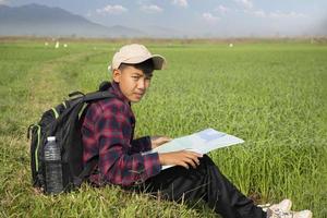 asiatisch Junge tragen Plaid Hemd und ein Deckel Sitzung auf Grat von Reis Paddy Feld, halten ein Karte und ein Fernglas, lesen Karte Vor beobachten Vögel, Uhr 2.5 Rauch und Ackerland Grenzen, Sanft Fokus. foto