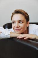 Frauen Lügen beim Zuhause auf das Couch Porträt mit ein kurz Haarschnitt im ein Weiß Shirt, lächeln, Depression im Jugendliche, Zuhause Urlaub foto