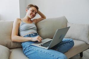 Teenager Mädchen Freiberufler mit Laptop Sitzung auf Couch beim Zuhause lächelnd im Zuhause Kleider und Brille mit kurz Haarschnitt, Lebensstil mit Nein Filter, kostenlos Kopieren Raum foto