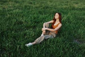 das rothaarig Frau sitzt im das Park auf das Grün Gras tragen ein Orange Spitze, Grün Hose, und Turnschuhe und sieht aus aus beim das Rahmen Sommer- Sonne foto