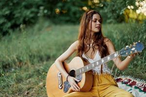 Mädchen Hippie Frau spielen Gitarre im umweltfreundlich Kleider Sitzung auf das Boden draußen im Natur im das fallen Aufpassen das Sonnenuntergang foto