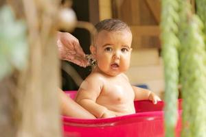 Nahansicht von ein Baby Mädchen suchen beim das Kamera, während ihr Mutter badet ihr im ein Becken im das Hinterhof. foto