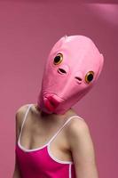 Frau tragen ein Rosa Fisch Kopf Halloween Maske steht und sieht aus beim das Kamera gegen ein Rosa Hintergrund foto