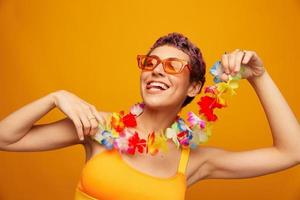 Porträt von ein Frau mit ein Blumen- hawaiisch Kranz um ihr Hals ist haben Spaß Tanzen und lächelnd im hell Kleider auf ein Orange Hintergrund tragen Sonnenbrille, Lebensstil Party hawaiisch Stil foto