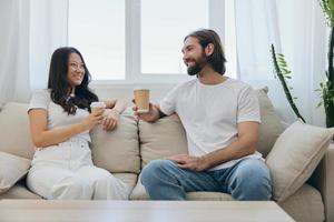 Mann und Frau Sitzung beim Zuhause auf das Couch im Weiß stilvoll T-Shirts Trinken Kaffee aus von Krabbe Tassen von ein Kaffee Geschäft und haben Spaß chatten lächelt und Lachen beim heim. männlich und weiblich Freundschaft foto