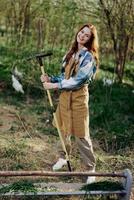 ein Frau Farmer im Arbeit Kleider und ein Schürze funktioniert draußen im Natur und hält ein Rechen zu versammeln Gras foto