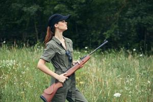 Frau auf draussen halten ein Gewehr im seine Hände schwarz Deckel Grün Overall hoch Gras Waffen foto
