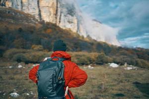 ein Frau im ein rot Jacke mit ein Rucksack und im ein warm Hut Spaziergänge auf das Gras im das fallen im das Berge foto