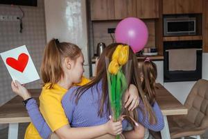 Töchter gratulieren ihr Mama auf Mutter Tag, ein Karte mit ein Herz, Blumen und ein Ballon beim Zuhause im das Küche, Sie alle Umarmung. Kinder Überraschung ihr Mutter zum das Urlaub. foto