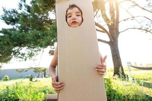 das Kind ist komisch Tanzen und täuschen um im ein Box Kostüm - - runden ausgeschnitten zum Gesicht und Hände. ziehen um zu ein Neu Haus, Kinder- Spiele von improvisiert bedeutet, ein passen mit Ihre besitzen Hände foto