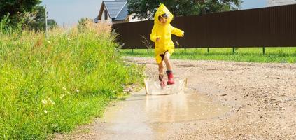 ein Mädchen im rot Gummi Stiefel und ein Gelb Regenjacke läuft durch Pfützen nach ein Regen im das Dorf. Sommer- Zeit, Freiheit, Kindheit foto