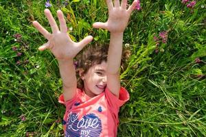 Porträt von ein Kind Mädchen im Sommer- Lügen im das Gras und Wildblumen mit Absätze und Palmen. Sommer- Zeit, Freiheit foto