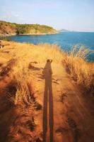 Frau nehmen Foto von selbst Porträt zum ihr Schatten im Seelandschaft im Sonnenuntergang und Meer Horizont mit Ruhe von Blau Himmel und trocken Gras Feld auf Berg von Kap im Phuket Insel, Thailand.