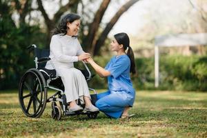 Alten asiatisch Senior Frau auf Rollstuhl mit asiatisch Vorsichtig Betreuer. Pflege- Zuhause Krankenhaus Garten Konzept. im Sonne Licht foto