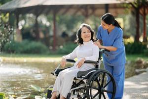 Alten asiatisch Senior Frau auf Rollstuhl mit asiatisch Vorsichtig Pflegekraft und ermutigen geduldig, Gehen im Garten. mit Pflege von ein Pflegekraft und Senior Gesundheit Versicherung. foto