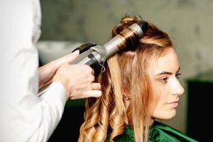 Friseur mit Eisstockschießen Zange Locken lange braun Haar auf das jung kaukasisch Mädchen im ein Schönheit Salon. foto