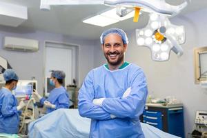 Porträt eines männlichen Chirurgen, der mit verschränkten Armen im Operationssaal des Krankenhauses steht. Teamchirurgen führen eine Operation durch, Arzt mittleren Alters schaut in die Kamera, in einem modernen Operationssaal foto
