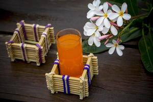 ein Glas von Orange Saft mit Wasser Glas Halter gemacht von Bambus Stöcke und Faser. dekorativ hölzern Tabelle mit schön Plumeria Blumen. Bambus Glas Halter. foto