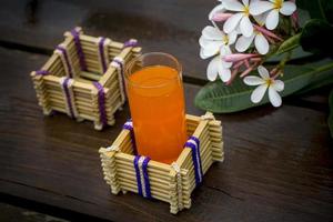 ein Glas von Orange Saft mit Wasser Glas Halter gemacht von Bambus Stöcke und Faser. dekorativ hölzern Tabelle mit schön Plumeria Blumen. Bambus Glas Halter. foto