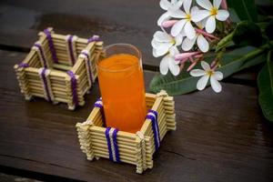 ein Glas von Orange Saft mit Wasser Glas Halter gemacht von Bambus Stöcke und Faser. dekorativ hölzern Tabelle mit schön Plumeria Blumen. Bambus Glas Halter. foto