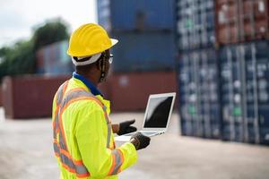 afrikanisch Techniker Dock Arbeiter im schützend Sicherheit Overall Uniform und mit Helm und verwenden Laptop Computer beim Ladung Container Versand Lagerhaus. Transport Import Export logistisch industriell foto